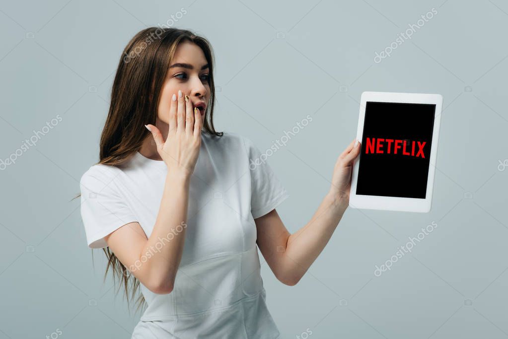 KYIV, UKRAINE - JUNE 6, 2019: shocked beautiful girl in white t-shirt showing digital tablet with Netflix app isolated on grey
