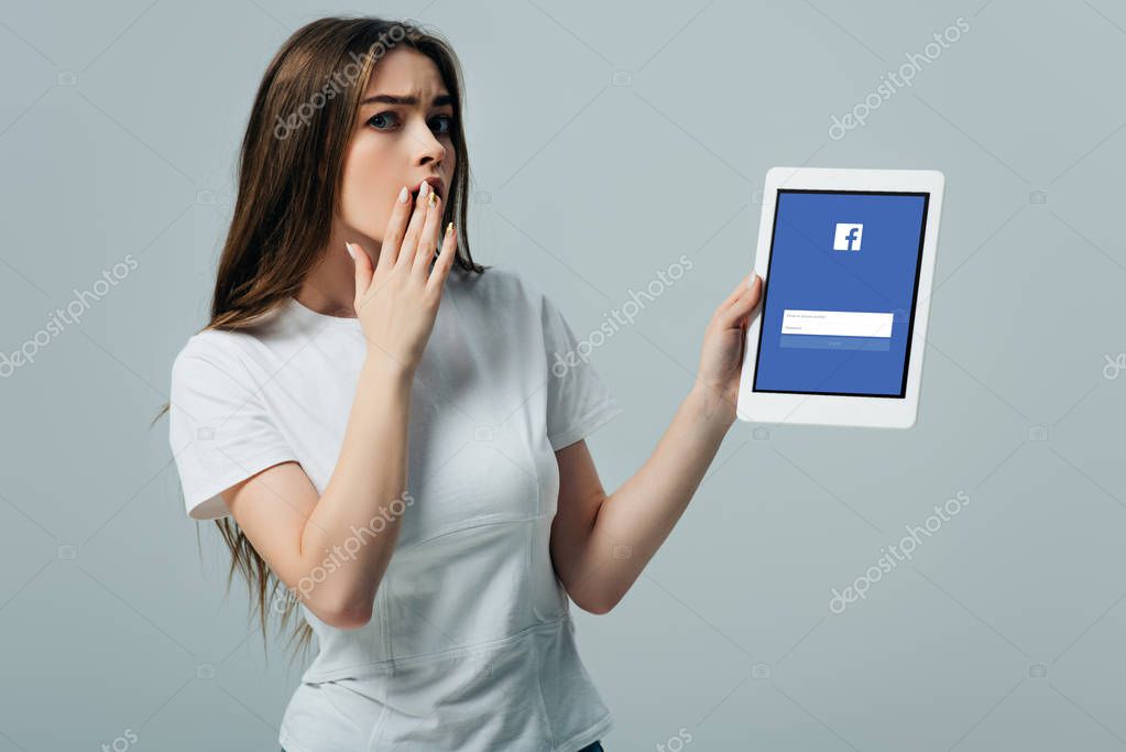 KYIV, UKRAINE - JUNE 6, 2019: shocked beautiful girl in white t-shirt showing digital tablet with facebook app isolated on grey