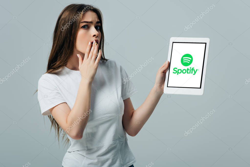 KYIV, UKRAINE - JUNE 6, 2019: shocked beautiful girl in white t-shirt showing digital tablet with Spotify app isolated on grey