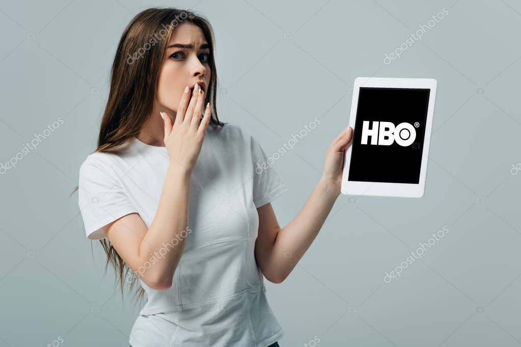 KYIV, UKRAINE - JUNE 6, 2019: shocked beautiful girl in white t-shirt showing digital tablet with HBO app isolated on grey