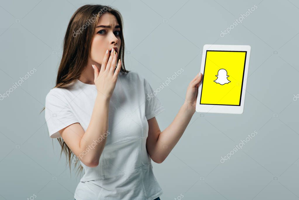 KYIV, UKRAINE - JUNE 6, 2019: shocked beautiful girl in white t-shirt showing digital tablet with Snapchat app isolated on grey