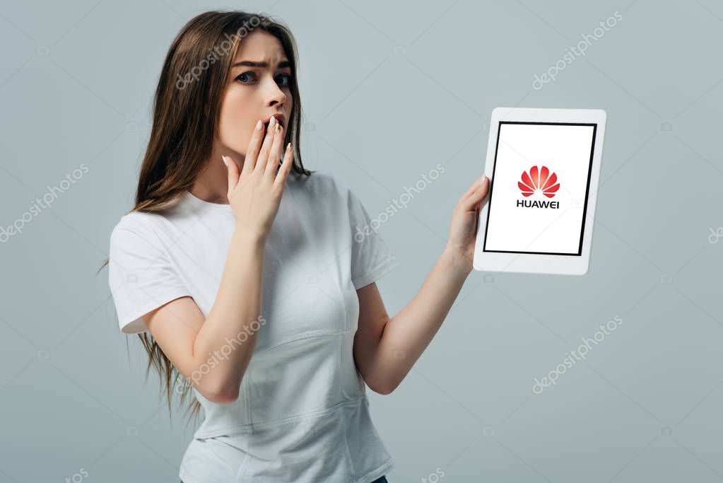 KYIV, UKRAINE - JUNE 6, 2019: shocked beautiful girl in white t-shirt showing digital tablet with Huawei icon isolated on grey