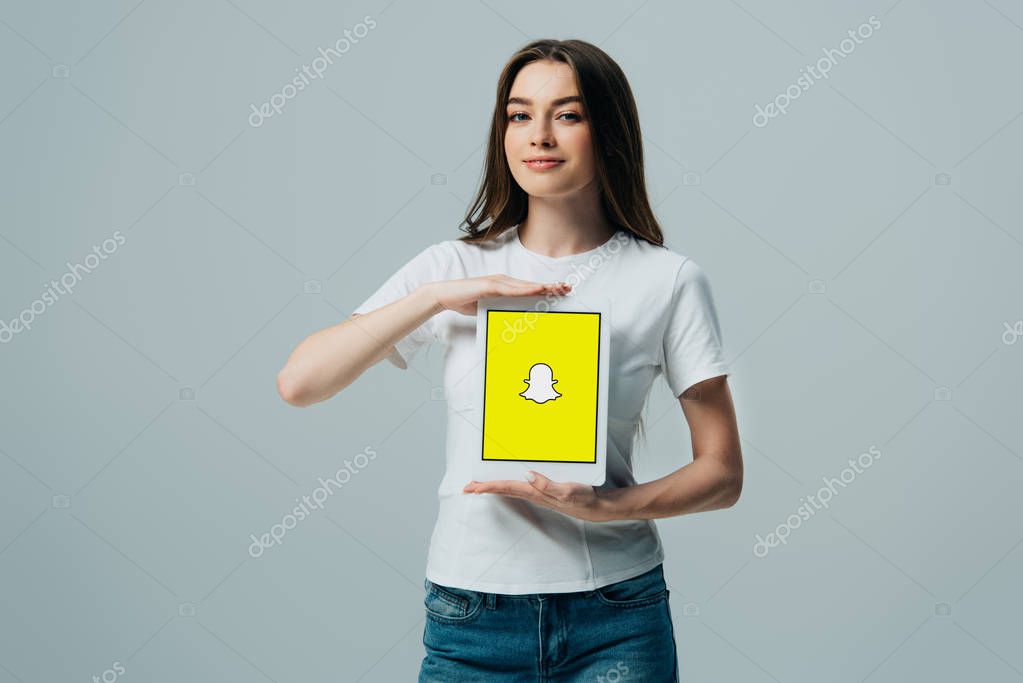 KYIV, UKRAINE - JUNE 6, 2019: smiling beautiful girl in white t-shirt showing digital tablet with Snapchat app isolated on grey