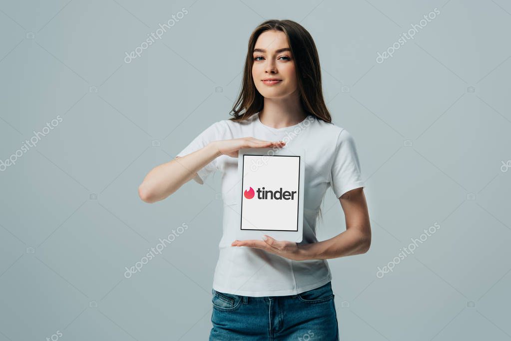 KYIV, UKRAINE - JUNE 6, 2019: smiling beautiful girl in white t-shirt showing digital tablet with Tinder app isolated on grey