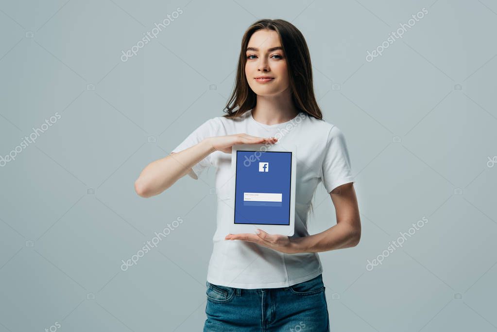 KYIV, UKRAINE - JUNE 6, 2019: smiling beautiful girl in white t-shirt showing digital tablet with Facebook app isolated on grey