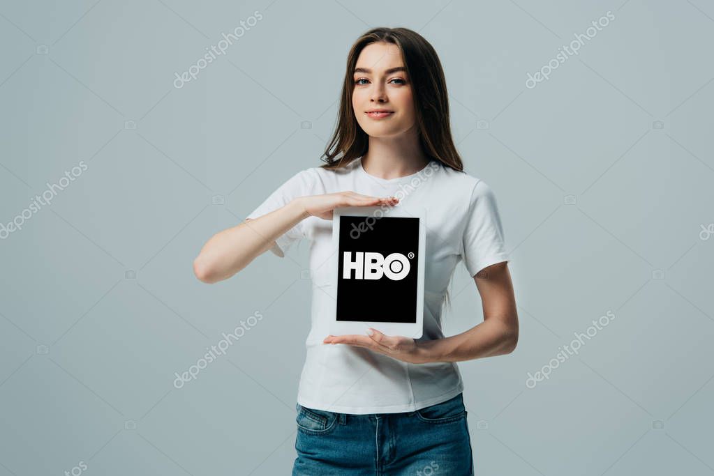 KYIV, UKRAINE - JUNE 6, 2019: smiling beautiful girl in white t-shirt showing digital tablet with HBO app isolated on grey