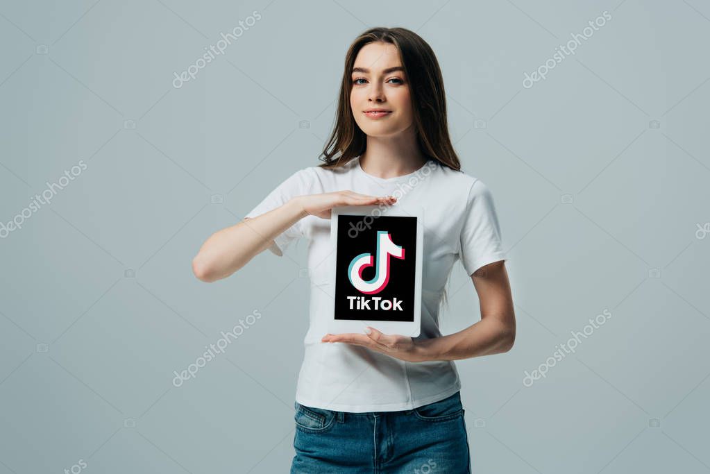 KYIV, UKRAINE - JUNE 6, 2019: smiling beautiful girl in white t-shirt showing digital tablet with Tik Tok app isolated on grey