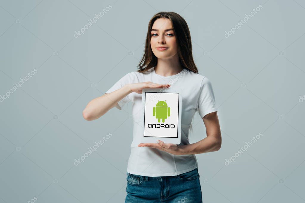 KYIV, UKRAINE - JUNE 6, 2019: smiling beautiful girl in white t-shirt showing digital tablet with Android icon isolated on grey