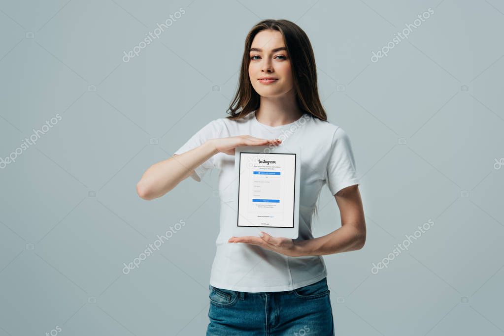 KYIV, UKRAINE - JUNE 6, 2019: smiling beautiful girl in white t-shirt showing digital tablet with Instagram app isolated on grey
