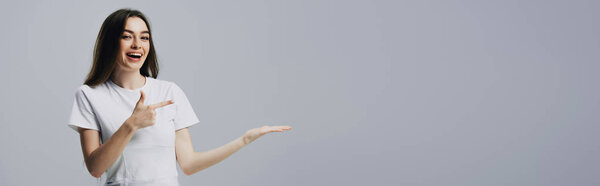 счастливая симпатичная девушка, показывающая пальцем и рукой на сером, панорамном снимке
