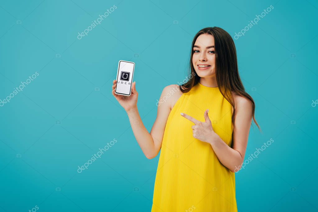 KYIV, UKRAINE - JUNE 6, 2019: happy beautiful girl in yellow dress pointing with finger at smartphone with uber app isolated on turquoise