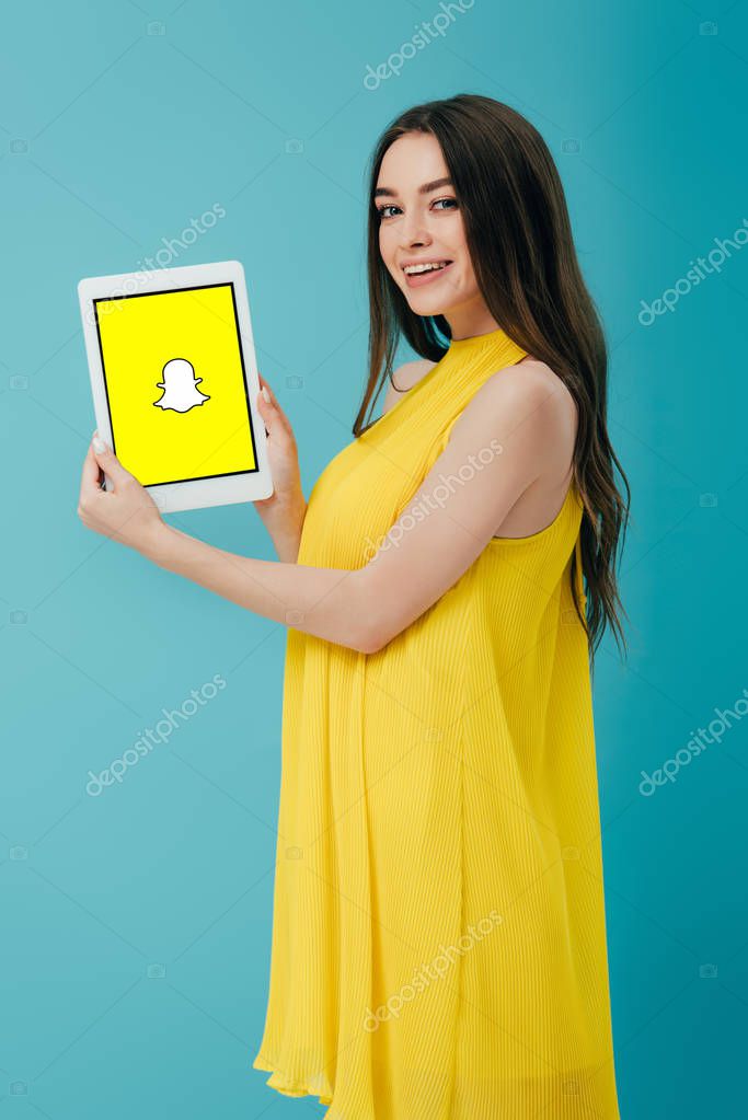 KYIV, UKRAINE - JUNE 6, 2019: smiling beautiful girl in yellow dress showing digital tablet with snapchat app isolated on turquoise