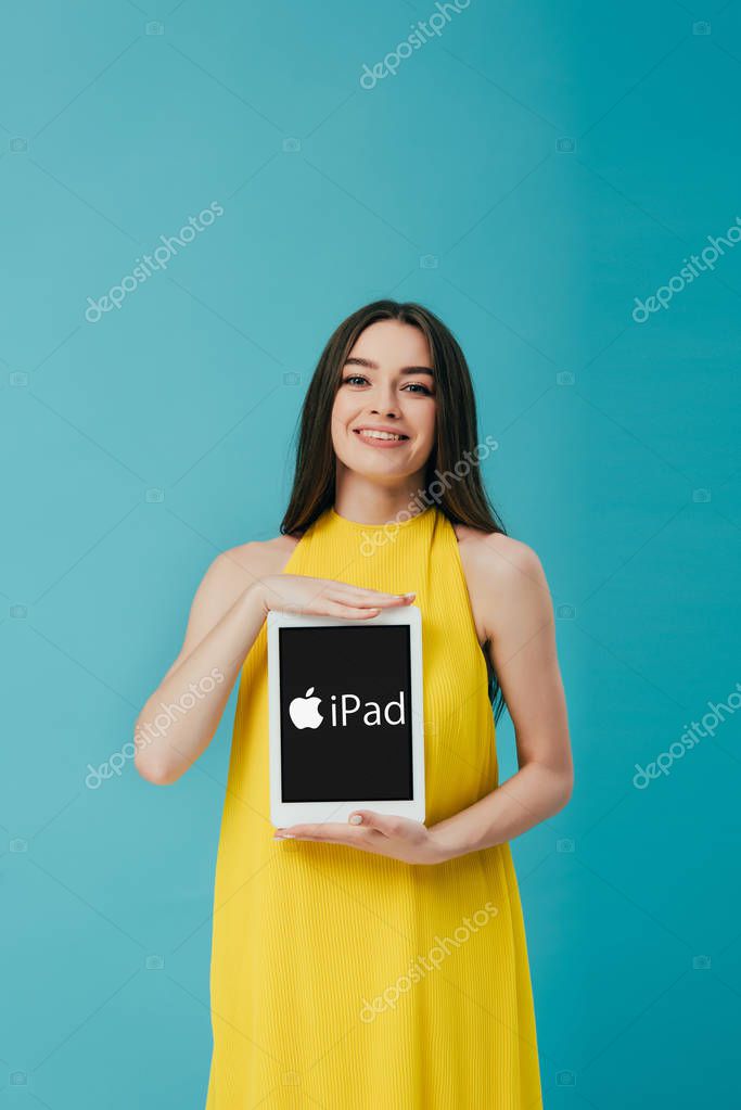 KYIV, UKRAINE - JUNE 6, 2019: smiling beautiful girl in yellow dress showing digital tablet with iPad logo isolated on turquoise