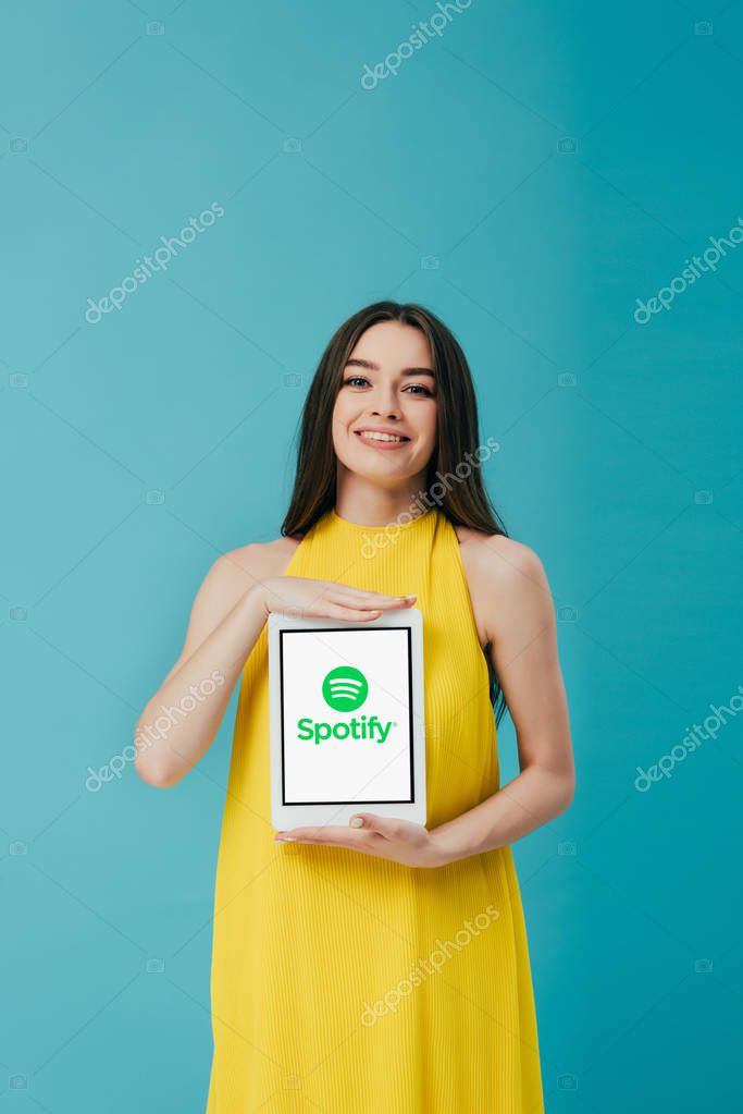 KYIV, UKRAINE - JUNE 6, 2019: smiling beautiful girl in yellow dress showing digital tablet with spotify app isolated on turquoise