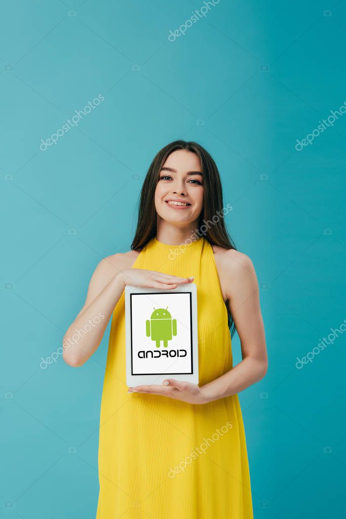 KYIV, UKRAINE - JUNE 6, 2019: smiling beautiful girl in yellow dress showing digital tablet with android logo isolated on turquoise