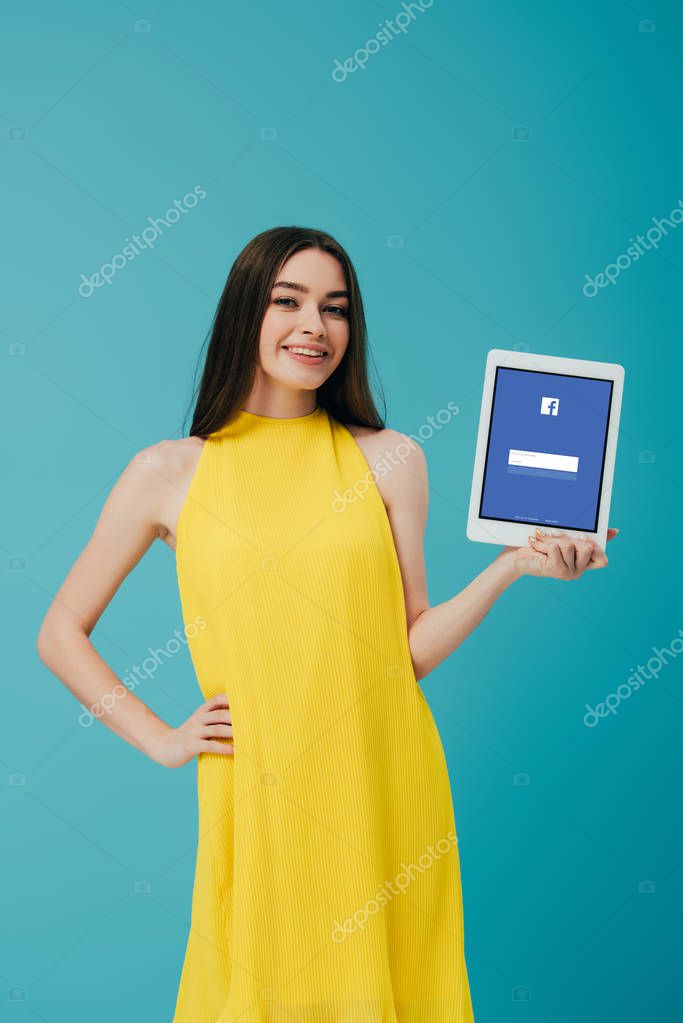KYIV, UKRAINE - JUNE 6, 2019: smiling girl in yellow dress with hand on hip showing digital tablet with facebook app isolated on turquoise