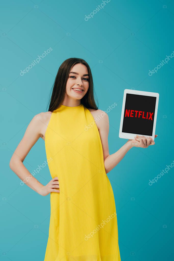 KYIV, UKRAINE - JUNE 6, 2019: smiling girl in yellow dress with hand on hip showing digital tablet with app isolated on turquoise
