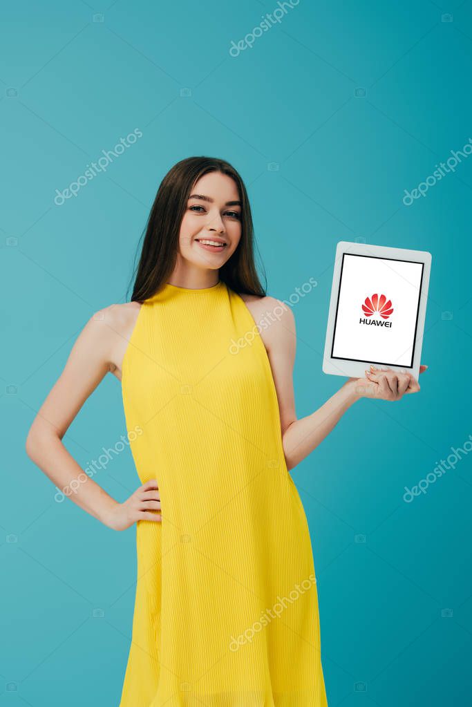 KYIV, UKRAINE - JUNE 6, 2019: smiling girl in yellow dress with hand on hip showing digital tablet with huawei logo isolated on turquoise