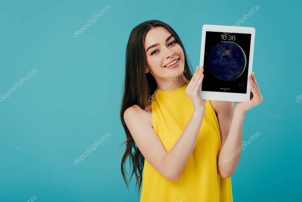 KYIV, UKRAINE - JUNE 6, 2019: happy beautiful girl in yellow dress showing digital tablet with apple lockscreen isolated on turquoise