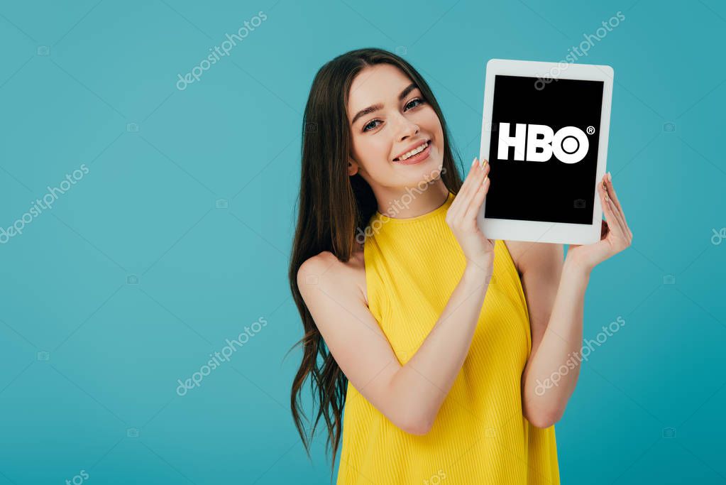 KYIV, UKRAINE - JUNE 6, 2019: happy beautiful girl in yellow dress showing digital tablet with HBO app isolated on turquoise