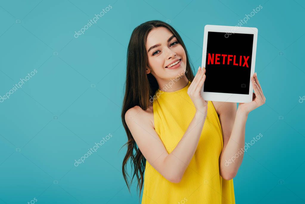 KYIV, UKRAINE - JUNE 6, 2019: happy beautiful girl in yellow dress showing digital tablet with Netflix app isolated on turquoise