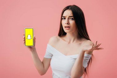 KYIV, UKRAINE - JUNE 6, 2019: beautiful confused girl holding smartphone with Snapchat app and showing shrug gesture isolated on pink clipart