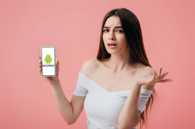 Kiev, Ukrayna - 6 Haziran 2019: Android logolu akıllı telefonu tutan ve pembe üzerinde izole omuz silkme jesti gösteren güzel kafası karışık kız