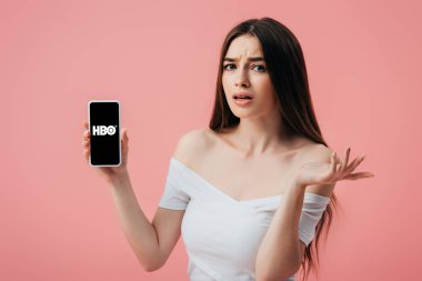 Kiev, Ukrayna - 6 Haziran 2019: Hbo uygulaması ile akıllı telefon tutan ve pembe izole omuz silkme jest gösteren güzel karışık kız