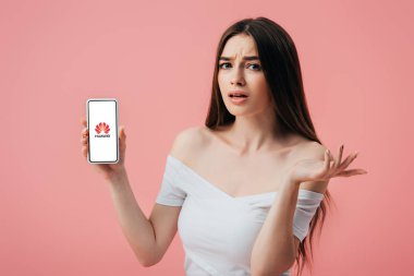 KYIV, UKRAINE - JUNE 6, 2019: beautiful confused girl holding smartphone with Huawei logo and showing shrug gesture isolated on pink clipart
