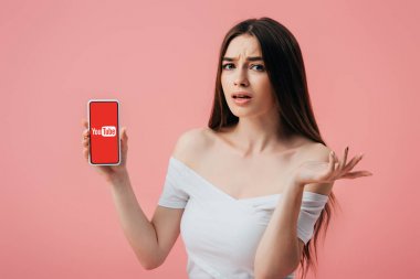 Kiev, Ukrayna - 6 Haziran 2019: youtube uygulaması ile akıllı telefon tutan ve pembe izole omuz silkme jest gösteren güzel karışık kız