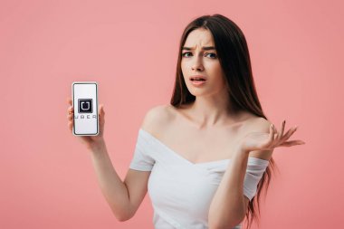 Kiev, Ukrayna - 6 Haziran 2019: Uber uygulaması ile akıllı telefon tutan ve pembe izole omuz silkme jest gösteren güzel şaşkın kız
