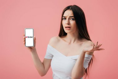 Kiev, Ukrayna - 6 Haziran 2019: tinder uygulaması ile akıllı telefon tutan ve pembe izole omuz silkme jest gösteren güzel karışık kız