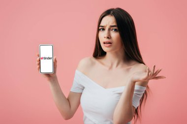 KYIV, UKRAINE - JUNE 6, 2019: beautiful confused girl holding smartphone with tinder app and showing shrug gesture isolated on pink clipart