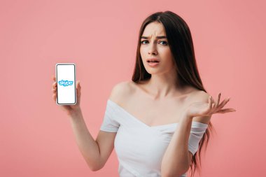 Kiev, Ukrayna - 6 Haziran 2019: Skype uygulaması ile akıllı telefon tutan ve pembe izole omuz silkme jest gösteren güzel şaşkın kız