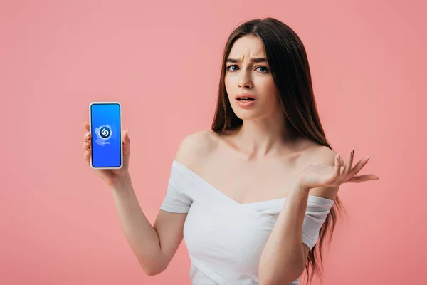 乌克兰 2019年6月6日 美丽的困惑的女孩拿着智能手机与Shazam应用程序 并显示耸肩手势孤立在粉红色 — 图库照片
