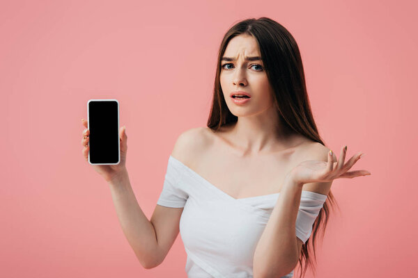 beautiful confused girl holding smartphone with blank screen and showing shrug gesture isolated on pink