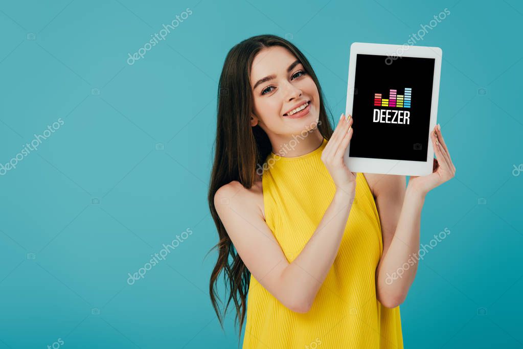 KYIV, UKRAINE - JUNE 6, 2019: happy beautiful girl in yellow dress showing digital tablet with deezer app isolated on turquoise