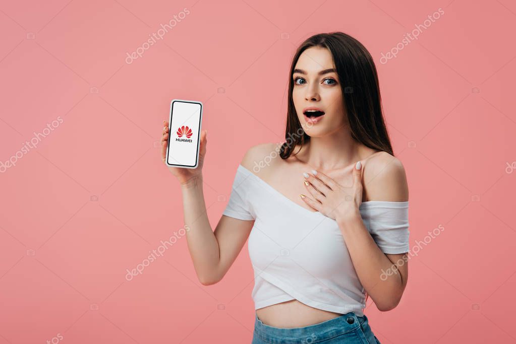KYIV, UKRAINE - JUNE 6, 2019: beautiful shocked girl holding smartphone with huawei logo isolated on pink