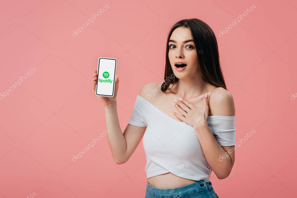 KYIV, UKRAINE - JUNE 6, 2019: beautiful shocked girl holding smartphone with Spotify app isolated on pink