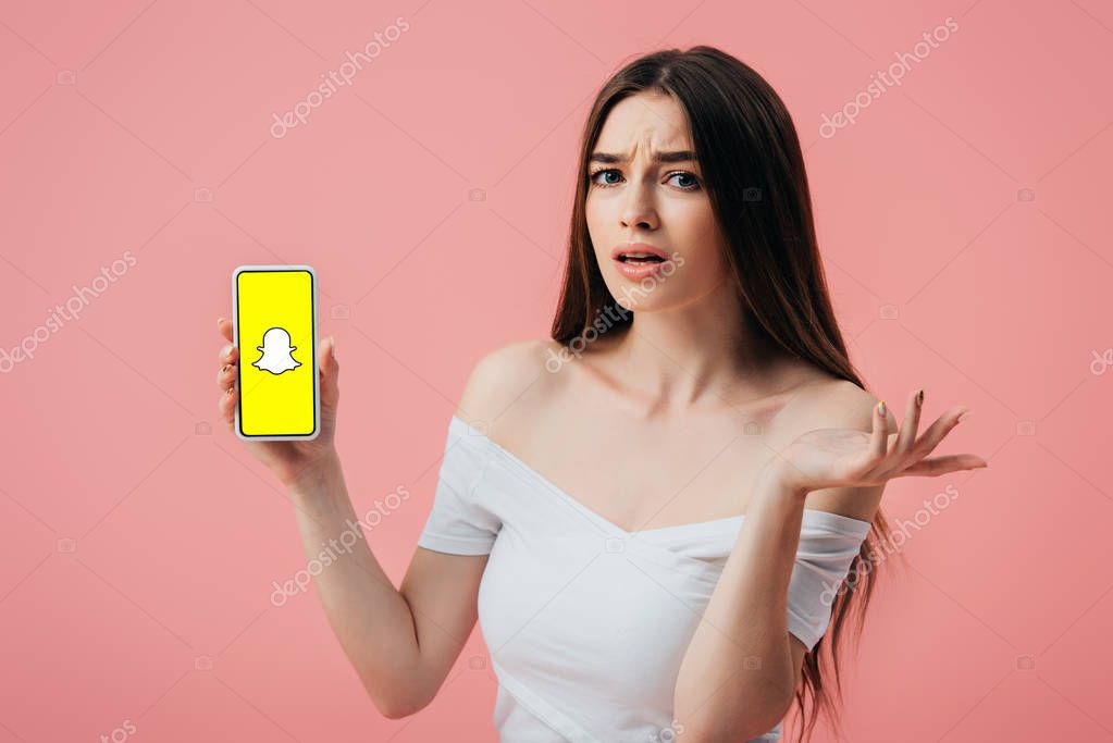 KYIV, UKRAINE - JUNE 6, 2019: beautiful confused girl holding smartphone with Snapchat app and showing shrug gesture isolated on pink