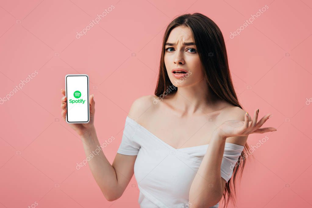KYIV, UKRAINE - JUNE 6, 2019: beautiful confused girl holding smartphone with Spotify app and showing shrug gesture isolated on pink