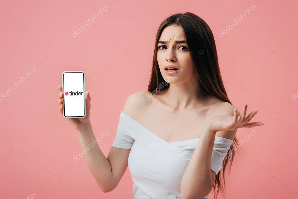 KYIV, UKRAINE - JUNE 6, 2019: beautiful confused girl holding smartphone with tinder app and showing shrug gesture isolated on pink