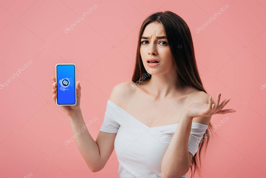 KYIV, UKRAINE - JUNE 6, 2019: beautiful confused girl holding smartphone with Shazam app and showing shrug gesture isolated on pink