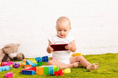 adorbale kid sitting among toys on green floor and holding digital device clipart