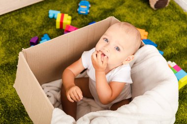Cute blue-eyed child sitting in cardboard box with white blanket on green floor clipart
