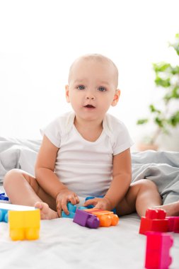 Funny barefoot baby in white clothes sitting on bed, playing with colorful construction and looking at camera clipart