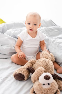 Beyaz elbiseli küçük çocuk yatakta oturan ve peluş ayı tutan