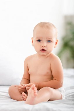 Cute barefoot child with blue eyes sitting on bed and looking at camera clipart