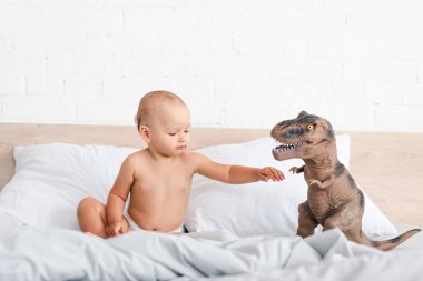 Sevimli küçük çocuk yatakta oturan ve kahverengi oyuncak dinozor el kaldırma