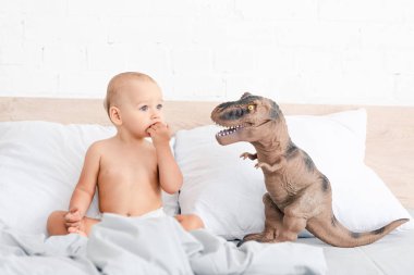 Kahverengi oyuncak dinozor yakınında hafif yatakta oturan küçük çocuk